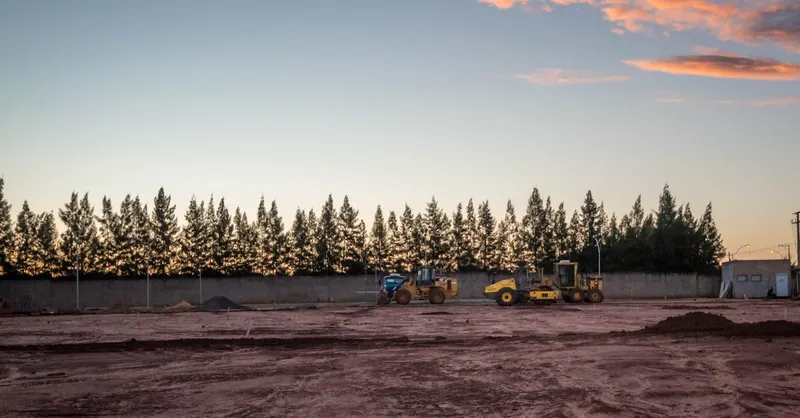 Heavy machinery on fenced site construction with forest under sky with clouds in evening time