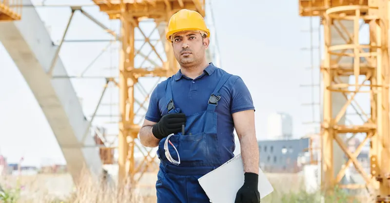 Man in safety gear holding blueprint on construction site with cranes.