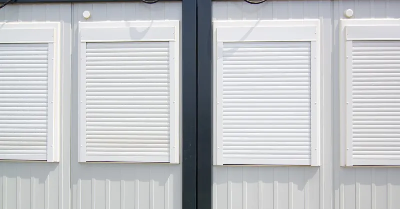 Close-up of a modular building with white shuttered windows and security features under a bright sky.