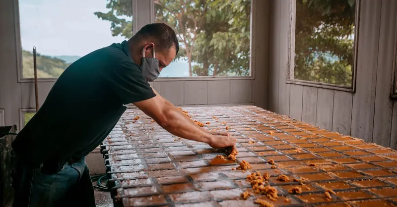 Man wearing mask, shaping clay bricks on workbench in indoor workshop.